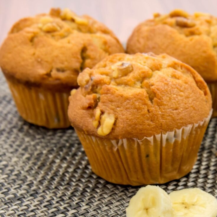 three banana bread muffins with nuts and sliced bananas on gray burlap