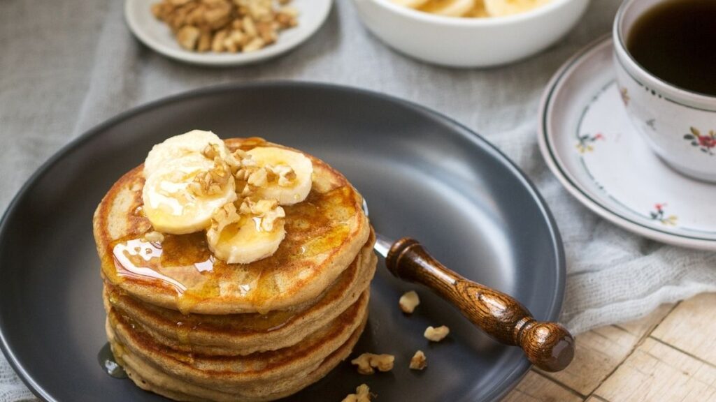 stack of whole wheat pancakes on gray plate topped with bananas and nuts