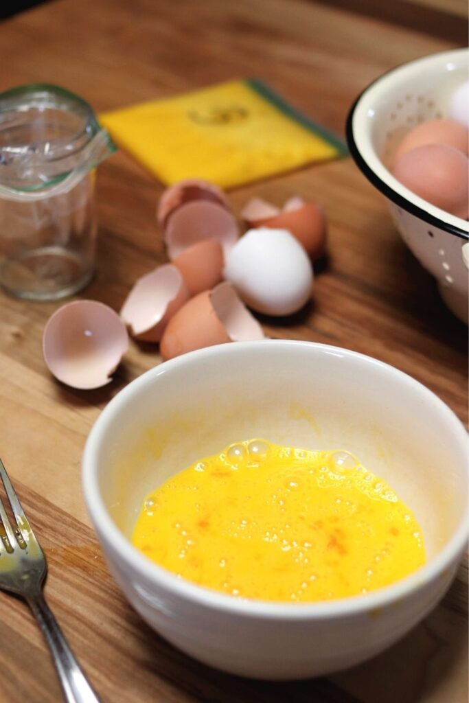 whisked eggs in a white bowl on a wooden table surrounded by egg shells, colander with farm fresh eggs, fork, mason jar lined with ziplock bag and a ziplock bag with whisked eggs for freezing